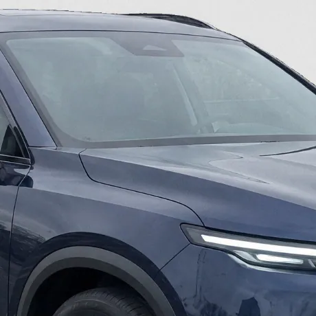 Close-up of the SUV’s windscreen and front wing in its real-photo studio image: the glass kept as photographed, pale with the lot’s grey sky, the cabin faint behind it.