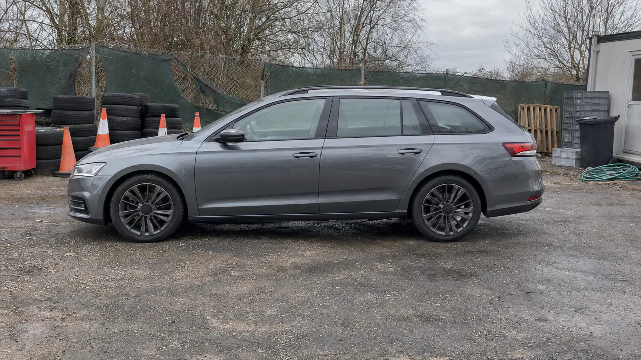 A fictional grey estate car in a cluttered corner of a car lot with tyres, cones and a fence behind it, side view; a sample photo made with TunePixel AI.