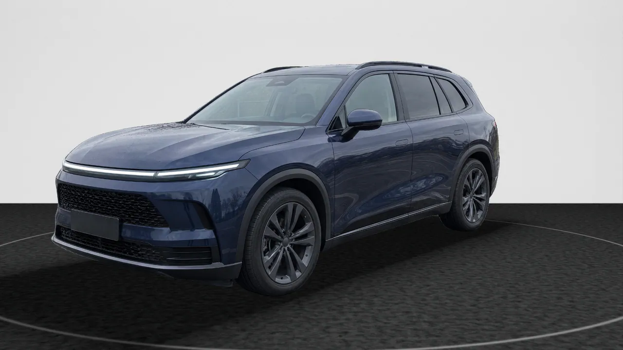 The fictional dark blue SUV, front three-quarter view from its other side, in the Black Turntable look: its lot photo’s car on dark charcoal carpet with a ring of light, in front of a white wall; a real-photo studio image.