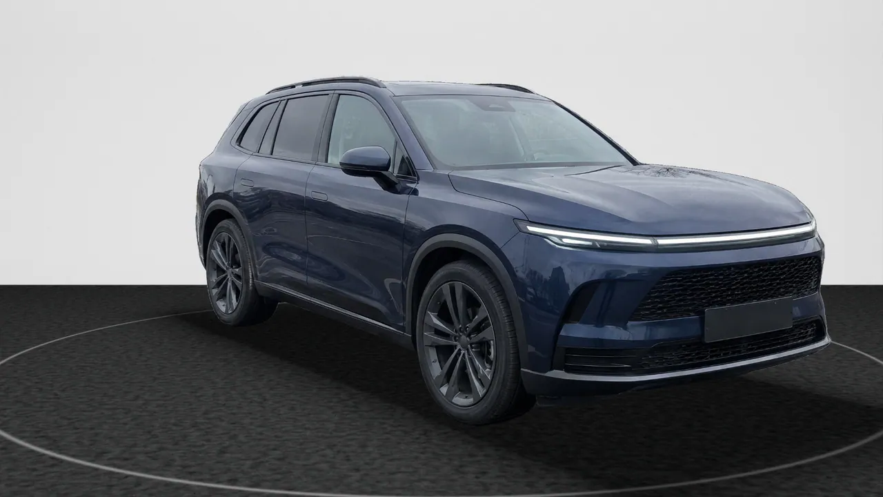 The fictional dark blue SUV, front three-quarter view, in the Black Turntable look: its lot photo’s car on dark charcoal carpet with a ring of light, in front of a white wall; a real-photo studio image.