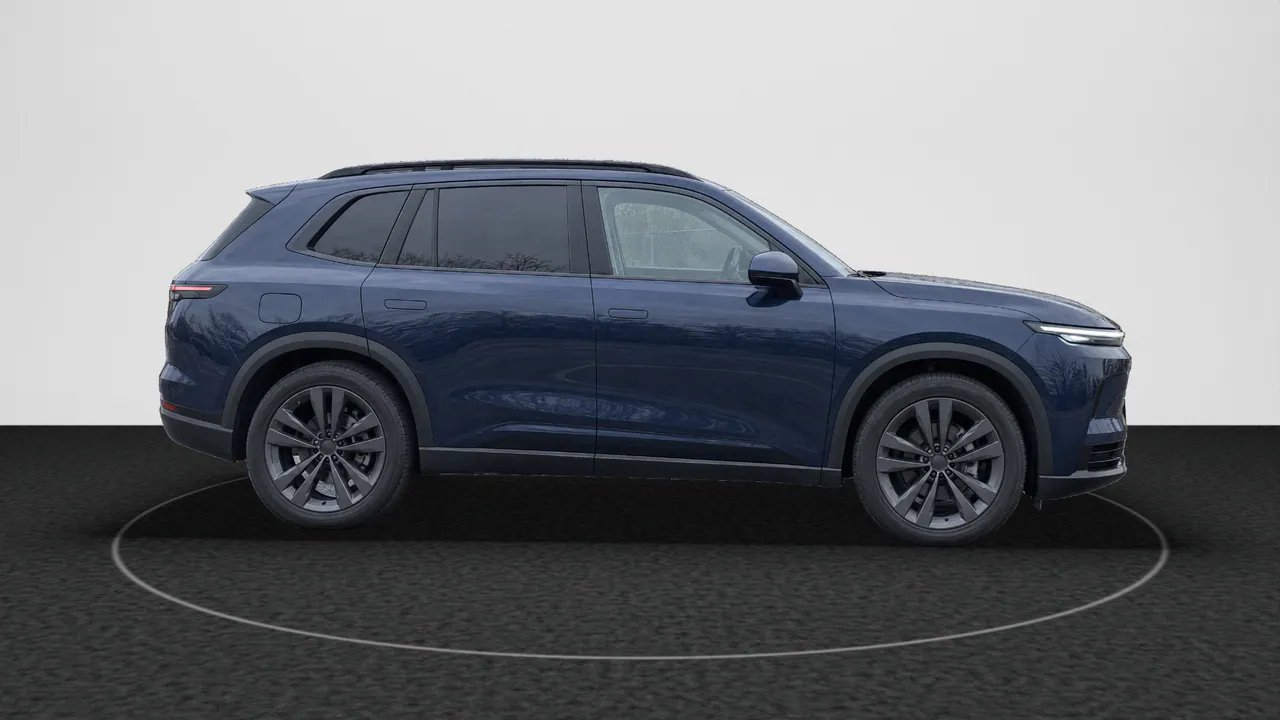 The fictional dark blue SUV, seen from the side, facing right, in the Black Turntable look: its lot photo’s car on dark charcoal carpet with a ring of light, in front of a white wall; a real-photo studio image.
